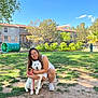 woman, dog, husky, white_dog, person_crouching, leash, park, grass, blue_sky, trees, residential_buildings, playground_tunnel, glasses, smile, sneakers, summer_dress, sunlight, shadow, portrait, outdoor