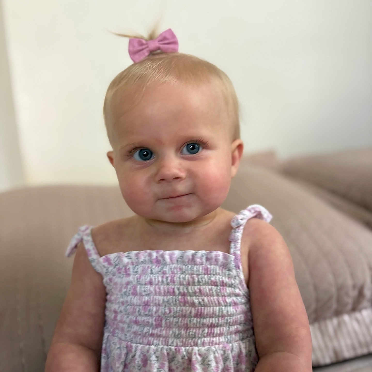 Huntleigh is registered to the contest to win money with this photo: baby, child, girl, blue_eyes, pink_bow, ponytail, floral_dress, sundress, cute, portrait, indoor, bedspread, quilted, soft_background, curious, smiling, infant, person, human, closeup