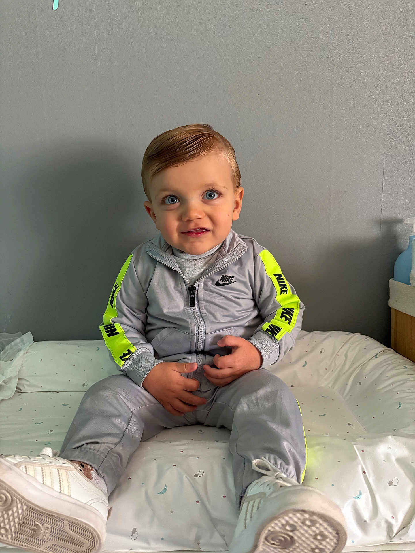 Lylio a rejoint le concours — aidez-le/la à gagner de superbes lots ! toddler, child, sitting, bed, tracksuit, nike, sneakers, blue_eyes, hair, gray_wall, indoor, casual_clothing, portrait, footwear, young_child, curious_expression, white_shoes, neon_stripes, hands, light