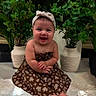 baby, barefoot, bow, child, cute, decor, dress, face, floor, floral_pattern, happy, indoor, infant, phone_screenshot, photograph, plants, portrait, potted_plants, sitting, smiling