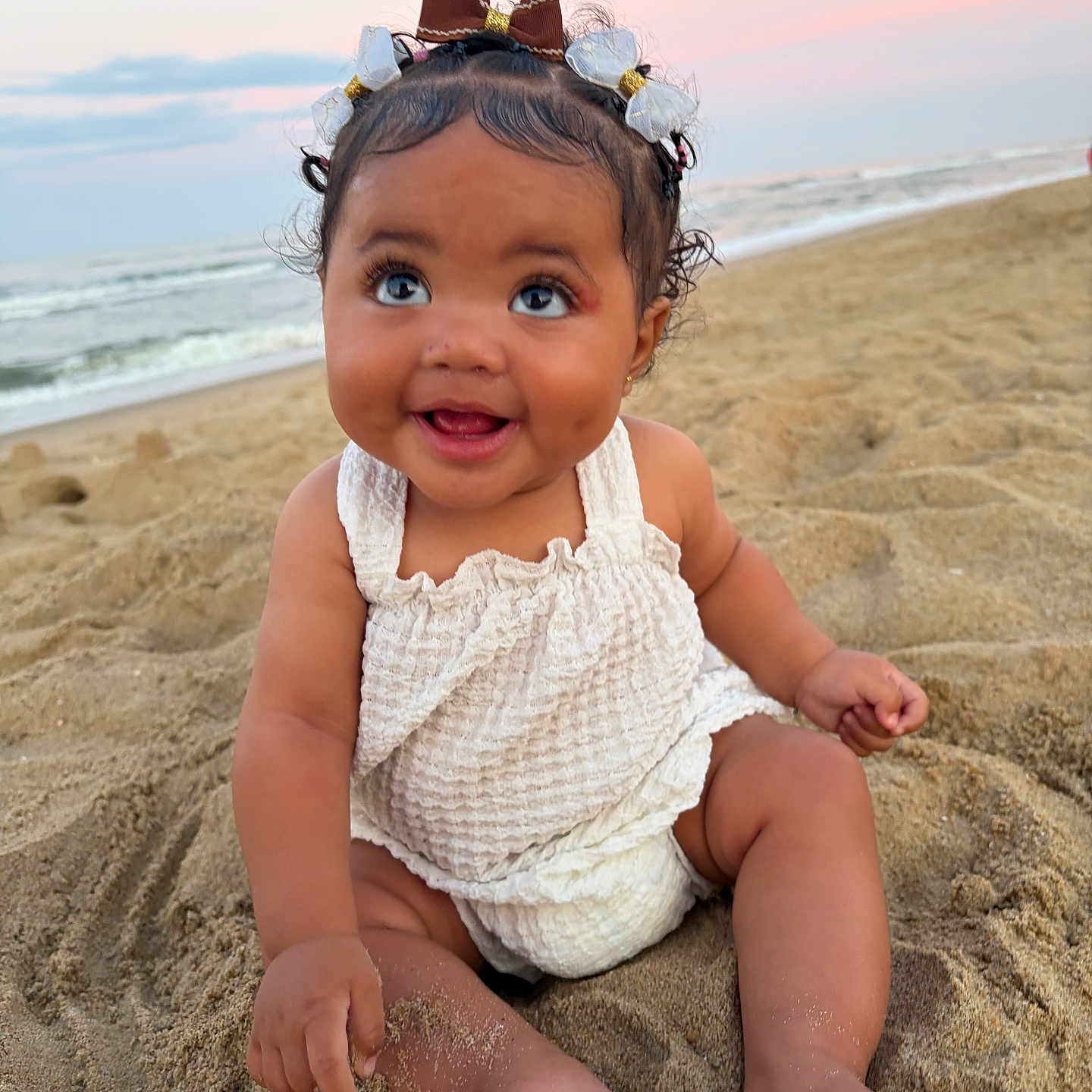 Adelina is registered to the contest to win money with this photo: baby, child, sand, beach, sunset, smile, happy, cute, bow, hair_accessory, ocean, water, sky, outdoor, summer, toddler, sitting, clothing, infant, portrait