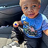 black_pants, blue_tshirt, car_interior, car_seat, casual_clothing, child, curious_expression, curly_hair, cute, daylight, holding_object, indoors, monster_truck, portrait, seatbelt, sitting, smiling, sneakers, toddler, young_child
