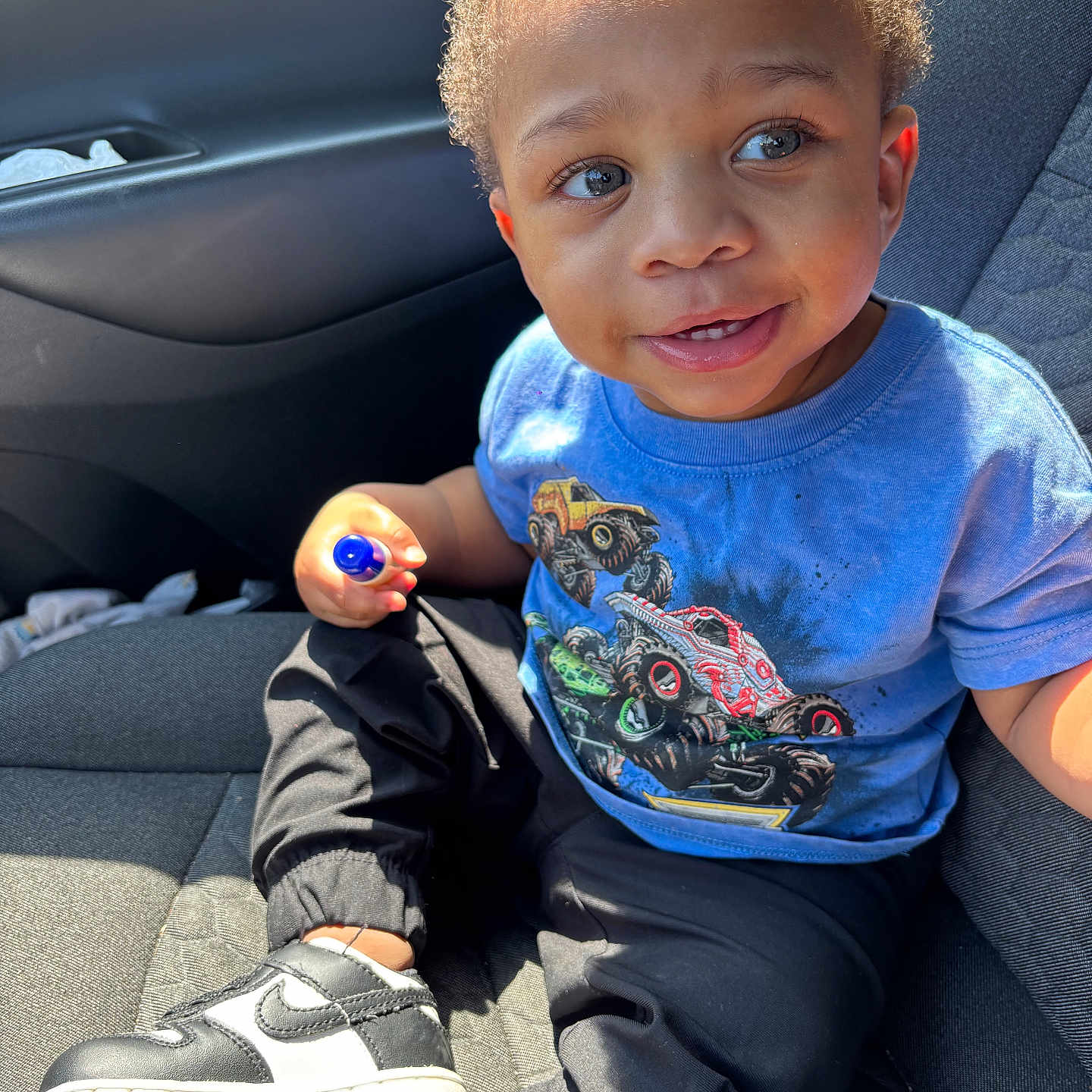 Drayden is registered to the contest to win money with this photo: black_pants, blue_tshirt, car_interior, car_seat, casual_clothing, child, curious_expression, curly_hair, cute, daylight, holding_object, indoors, monster_truck, portrait, seatbelt, sitting, smiling, sneakers, toddler, young_child