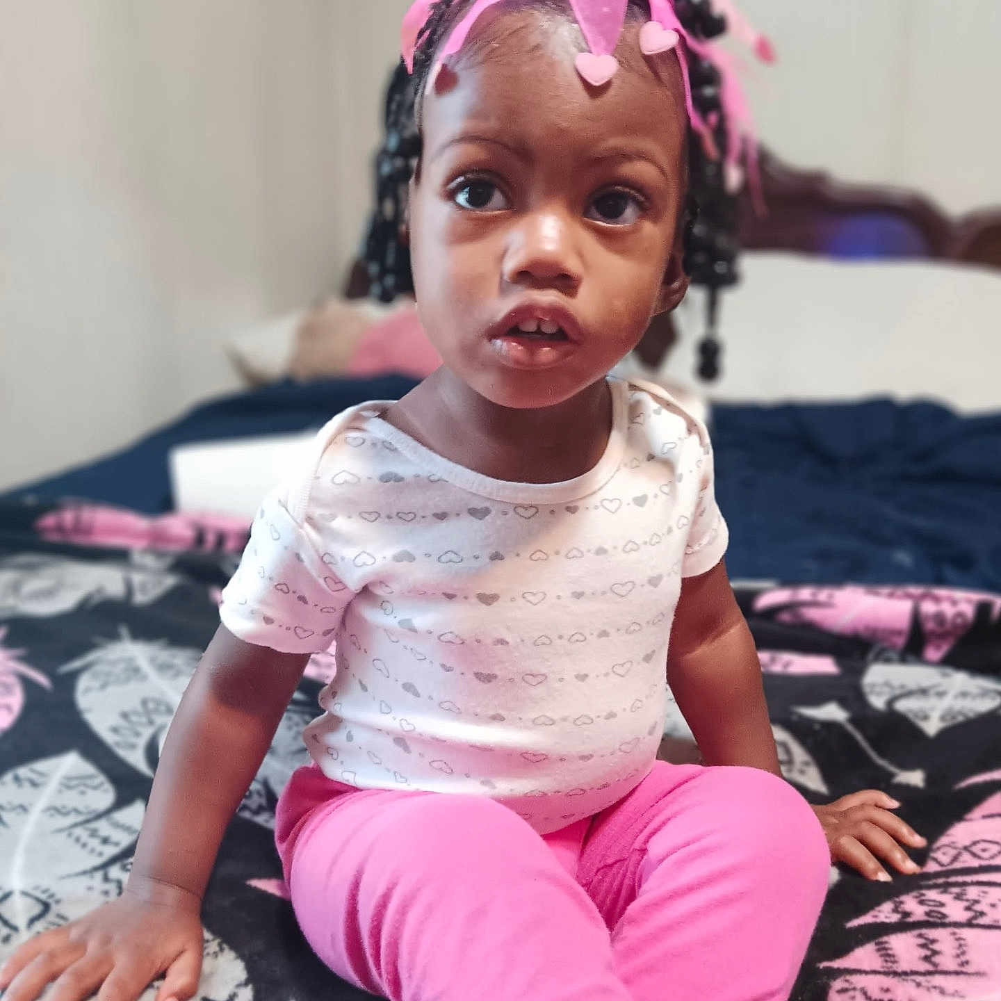 Mya joined the competition — help win amazing prizes! toddler, child, girl, pink_pants, pink_bow, headband, braided_hair, patterned_blanket, bedroom, indoor, sitting, curious, clothing, person, young_child, portrait, expression, hand, furniture, casual