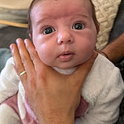 Kiara a rejoint le concours — aidez-le/la à gagner de superbes lots ! baby, infant, newborn, face, wide_eyes, hand, adult_hand, ring, onesie, blanket, pillow, couch, close_up, portrait, indoors, soft_texture, gaze, eyelashes, support, cozy