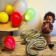 Jaxon is registered to the contest to win money with this photo: balloons, celebration, child, clapping, curly_hair, green_balloon, happy, indoor, party, person, portrait, red_balloon, shorts, silver_balloon, smiling, socks, toddler, white_balloon, wooden_table, yellow_balloon