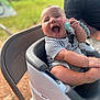 baby, high_chair, pacifier, striped_clothing, outdoor, greenery, happy, laughing, child, person, infant, seat, smile, holding, arm, casual_clothing, blurred_background, sunlight, cheerful, playful