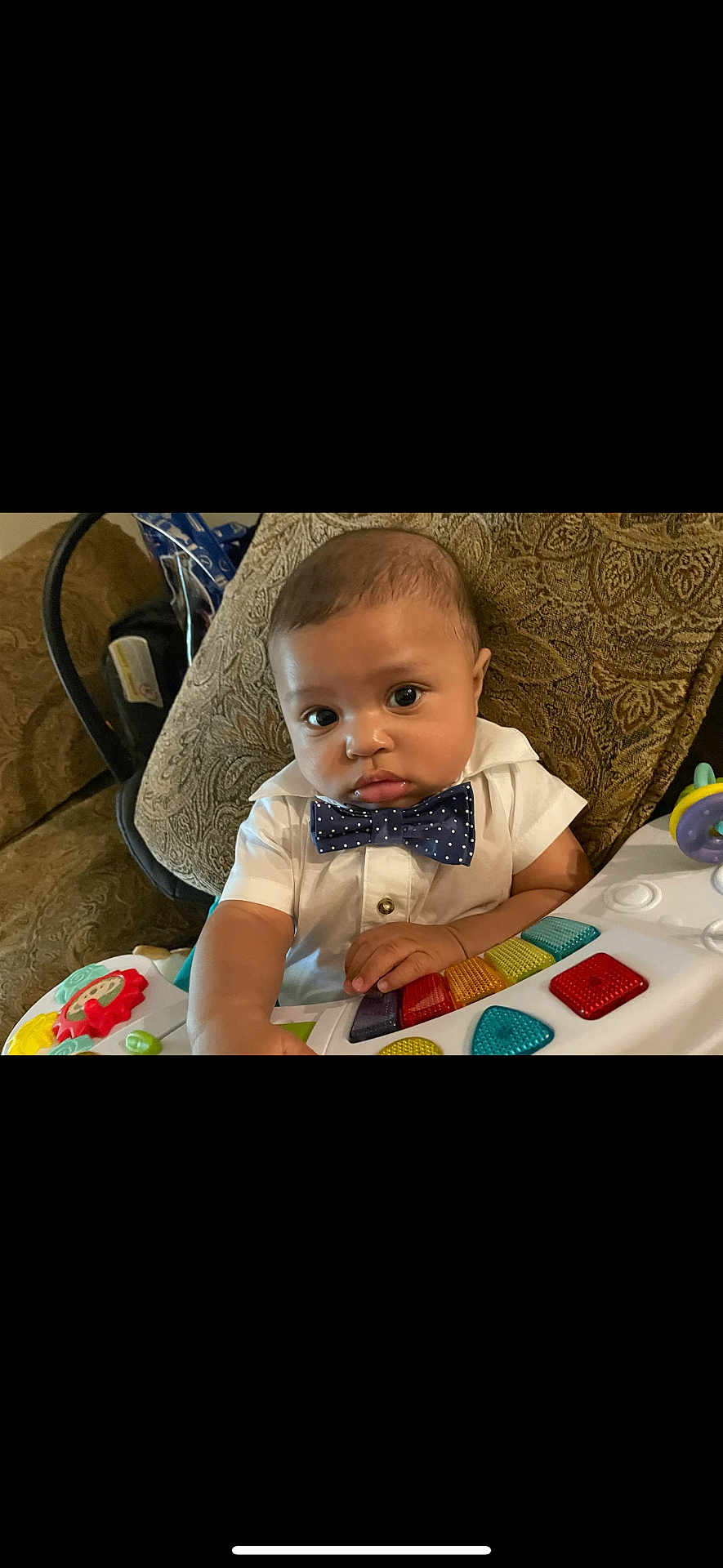 Dawson is registered to the contest to win money with this photo: baby, child, bow_tie, white_shirt, toy, colorful, piano_toy, cushion, patterned_fabric, indoor, furniture, person, infant, closeup, face, hands, seated, expression, cute, play