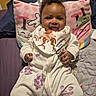 Ariyana is registered to the contest to win money with this photo: baby, bed, bib, blanket, cartoon, child, clothing, cozy, cute, face, hands, happy, indoor, infant, onesie, person, pillow, portrait, seated, smiling