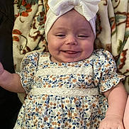 Esme is registered to the contest to win money with this photo: baby, smiling, headband, bow, floral_dress, blanket, pattern, cute, infant, child, happy, lying_down, small_hand, soft_texture, indoors, closeup, person, face, apparel, comfort