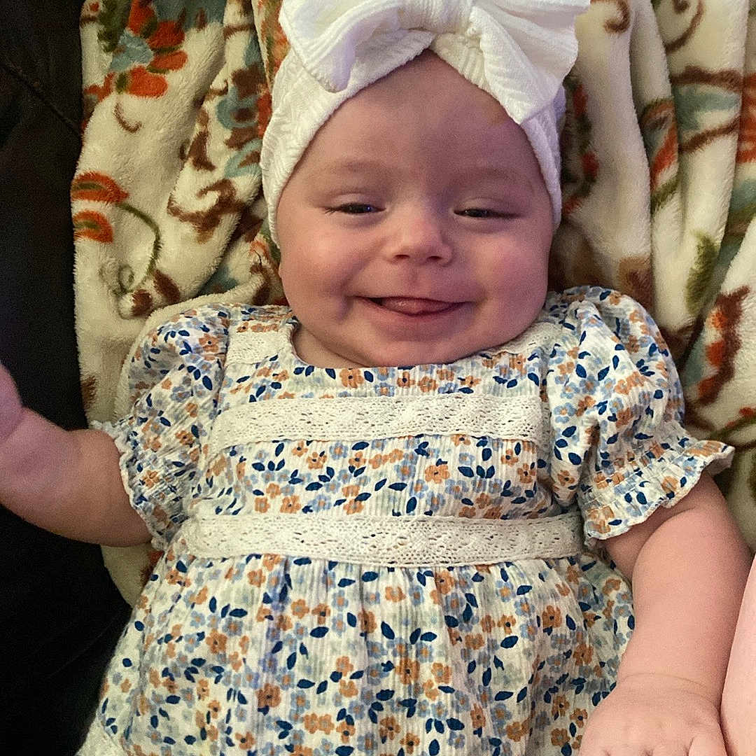 Esme is registered to the contest to win money with this photo: baby, smiling, headband, bow, floral_dress, blanket, pattern, cute, infant, child, happy, lying_down, small_hand, soft_texture, indoors, closeup, person, face, apparel, comfort