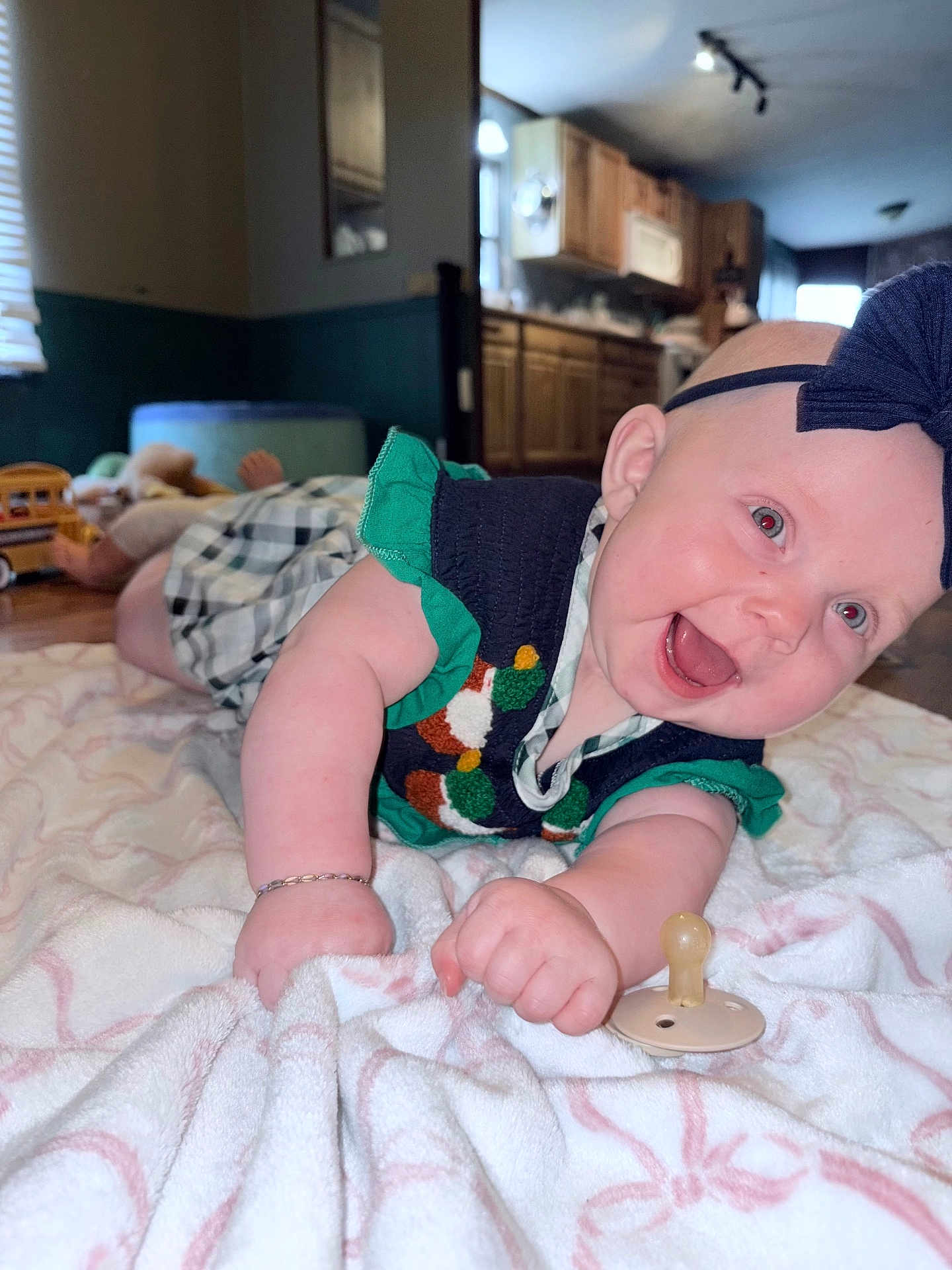 Laiken joined the competition — help win amazing prizes! baby, smiling, infant, blanket, headband, dress, pacifier, indoors, floor, wooden_floor, kitchen, cabinet, toy, child, happy, lying_down, bracelet, closeup, person, home