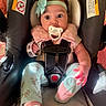 baby, pacifier, car_seat, headband, bow, pink_neck_pillow, socks, pants, infant, child, seatbelt, straps, indoors, cute, wide_eyes, person, baby_clothes, sitting, soft_lighting, comfort