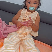 Isabelle a rejoint le concours — aidez-le/la à gagner de superbes lots ! toddler, child, pacifier, yellow_bows, curly_hair, striped_outfit, pink_blanket, gray_couch, indoor, sitting, cute, portrait, baby, clothing, person, face, hand, feet, looking, young_child