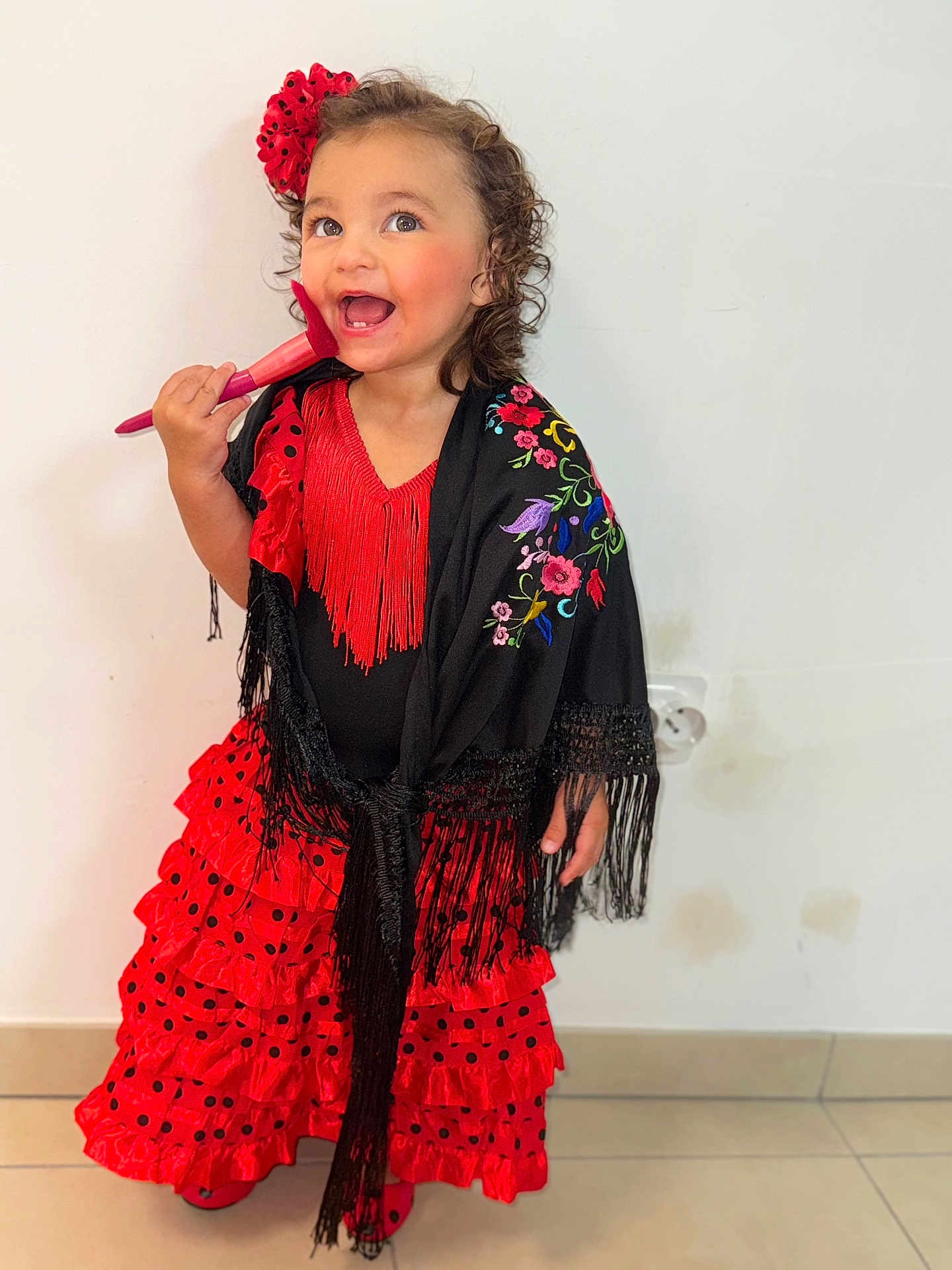 Isabelle a rejoint le concours — aidez-le/la à gagner de superbes lots ! child, girl, flamenco_dress, red_dress, black_shawl, flower_hair_accessory, curly_hair, makeup_brush, smiling, happy, standing, indoor, tile_floor, white_wall, colorful_embroidery, frills, polka_dots, portrait, playful, fashion