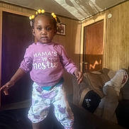 Kamorah joined the competition — help win amazing prizes! toddler, child, indoor, clothing, purple_hoodie, floral_pants, crocs, yellow_beads, hair_accessories, couch, wooden_paneling, shadow, person_in_background, living_room, smiling, standing, portrait, home, casual, cute