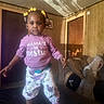 toddler, child, indoor, clothing, purple_hoodie, floral_pants, crocs, yellow_beads, hair_accessories, couch, wooden_paneling, shadow, person_in_background, living_room, smiling, standing, portrait, home, casual, cute