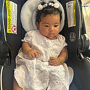 Nyla joined the competition — help win amazing prizes! baby, child, car_seat, white_dress, curly_hair, bows, tongue_out, infant, cute, portrait, sitting, indoors, black, fabric, seat_belt, warning_label, skin, hand, face, clothing