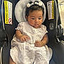 baby, child, car_seat, white_dress, curly_hair, bows, tongue_out, infant, cute, portrait, sitting, indoors, black, fabric, seat_belt, warning_label, skin, hand, face, clothing