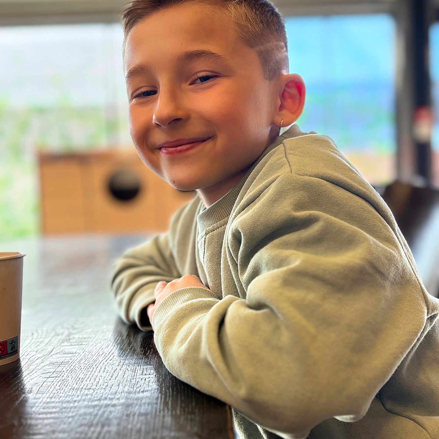 Tiago participe au concours pour gagner de l'argent avec cette photo : child, boy, smile, face, hairstyle, earring, sweatshirt, table, indoor, natural_light, portrait, casual, young, happy, person, wood, blurred_background, confident, seated, relaxed