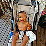 baby, stroller, smiling, happy, child, toy, wooden_deck, clothing, seatbelt, bare_chest, hand, foot, face, person, outdoor, sunlight, fabric, black_hair, infant, play