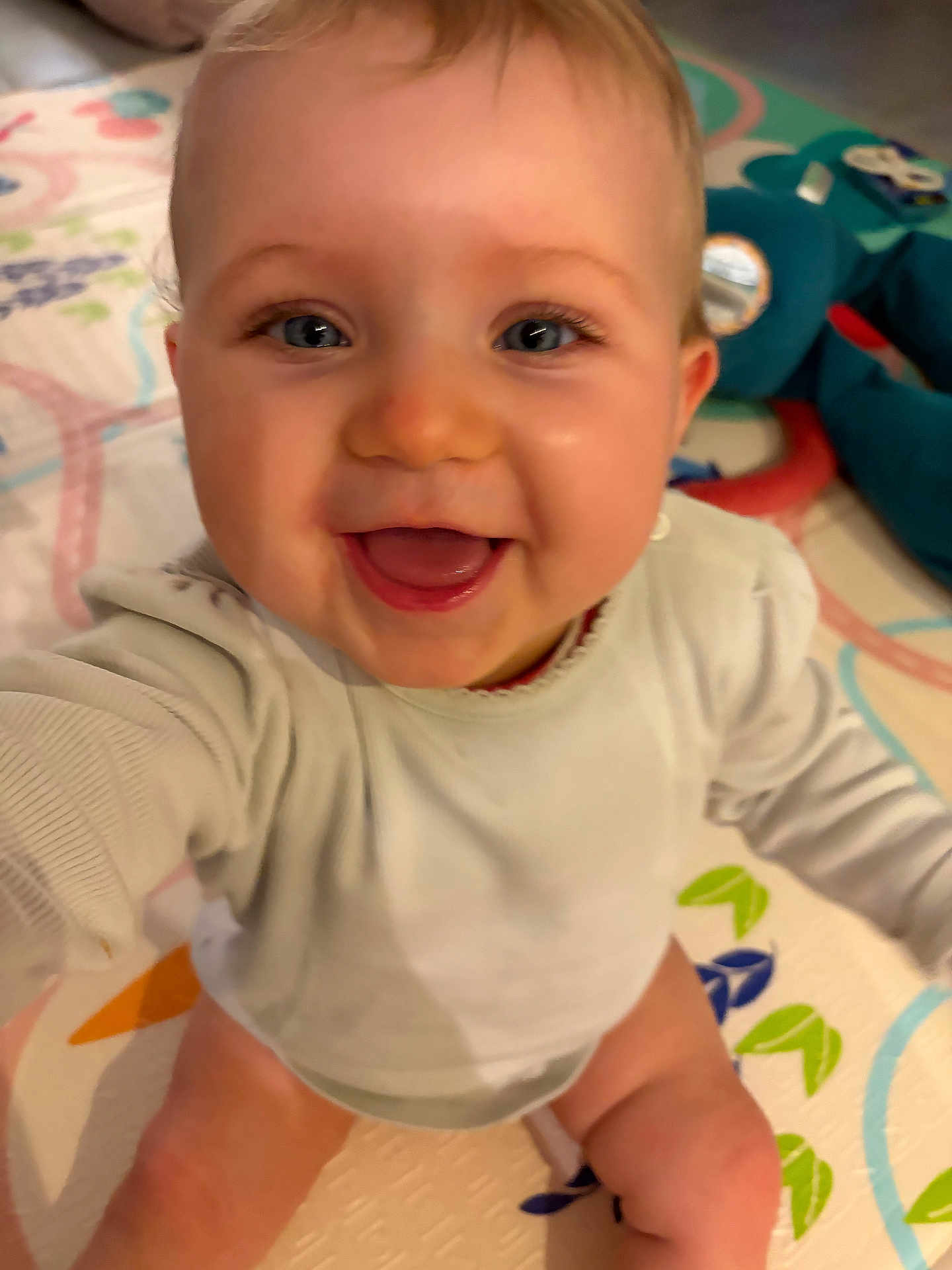 Adèle a rejoint le concours — aidez-le/la à gagner de superbes lots ! baby, infant, smiling, close_up, face, blue_eyes, eyelashes, chubby_cheeks, open_mouth, onesie, play_mat, toy, plush, sitting, hands, legs, home_interior, cozy, joyful, candid