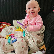 Dallas is registered to the contest to win money with this photo: baby, infant, smiling, pink_clothes, blanket, colorful_toy, teething_toy, couch, soft_throw, portrait, face, happy, sitting, indoor, cozy, playtime, floral_blanket, fur_texture, earring, cute