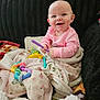 baby, infant, smiling, pink_clothes, blanket, colorful_toy, teething_toy, couch, soft_throw, portrait, face, happy, sitting, indoor, cozy, playtime, floral_blanket, fur_texture, earring, cute