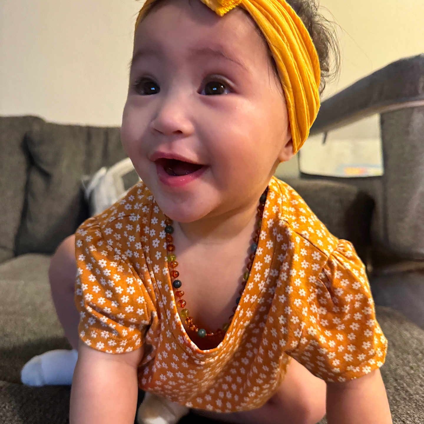 Bernadette is registered to the contest to win money with this photo: baby, beaded_necklace, bright, child, couch, crawling, cute, face, floral_dress, happy, headwear, home, indoors, infant, person, portrait, smiling, socks, yellow_headband, young_child