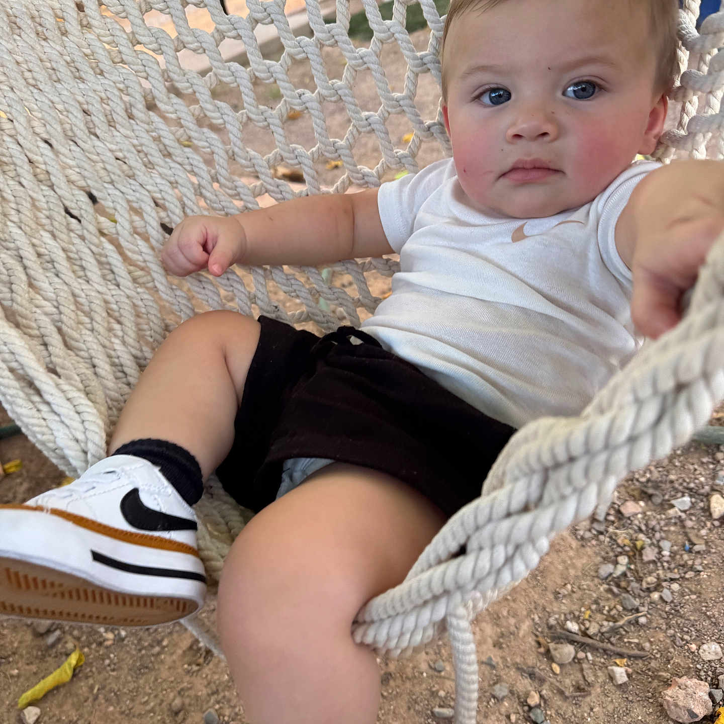 Zavian is registered to the contest to win money with this photo: baby, child, toddler, portrait, face, eyes, serious_expression, outdoor, hammock, rope, sitting, lying_down, white_shirt, black_shorts, sneakers, socks, playground, dirt, gravel, photography