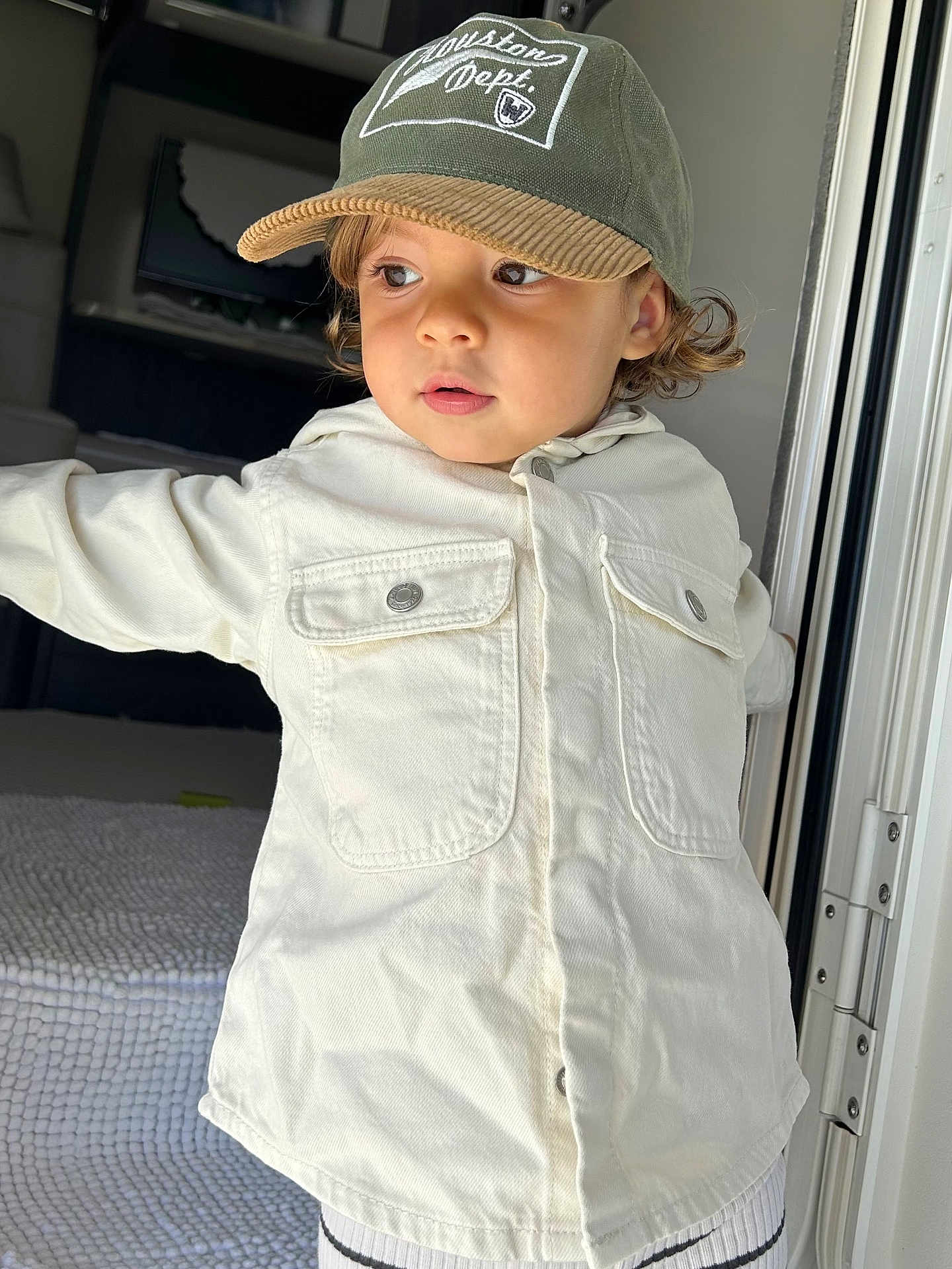 Elie participe au concours pour gagner de l'argent avec cette photo : toddler, child, cap, hat, curly_hair, jacket, white_jacket, indoor, door, natural_light, portrait, person, young_child, face, clothing, cute, curious, standing, expression, casual