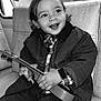 Elie participe au concours pour gagner de l'argent avec cette photo : child, toddler, guitar, smiling, black_and_white, jacket, plaid_shirt, seat, vehicle_interior, happy, portrait, music, hands, clothing, cute, young, indoor, hair, face, expressive