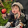 baby, child, stroller, apple, knitted_hat, knitted_sweater, socks, outdoor, greenery, sunlight, cute, happy, reaching, red, nature, infant, portrait, face, hand, seat