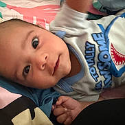 Javian is registered to the contest to win money with this photo: baby, bedding, blanket, blue, brown_hair, child, close_up, clothing, cozy, eyes, face, green, hand, indoors, infant, lying_down, pink, portrait, shark_graphic, smiling