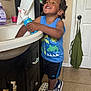 child, toddler, smiling, blue_shirt, black_shorts, bathroom, sink, step_stool, sneakers, nike_shoes, soap_dispenser, paper_towel, cabinet, tile_floor, door, towel, happy, indoor, person, braided_hair