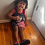 child, toddler, smiling, curly_hair, sitting, floor, wooden_floor, wall, door, cap, tank_top, shorts, flip_flops, indoors, happy, person, portrait, clothing, american_flag, red_white_blue