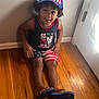 child, boy, smiling, sitting, hat, american_flag, shorts, tank_top, wooden_floor, indoor, door, curly_hair, casual_clothing, flip_flops, light, shadow, happy, patriotic, home, person