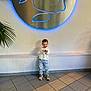 Hugo a rejoint le concours — aidez-le/la à gagner de superbes lots ! child, boy, smiling, standing, white_shirt, blue_jeans, sneakers, tile_floor, indoor, wall, plant, sign, blue_light, happy, casual_clothing, portrait, young, cute, fashion, playful