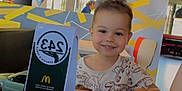Hugo participe au concours pour gagner de l'argent avec cette photo : child, toddler, smile, dinosaur_shirt, table, smartphone, hand, restaurant, fast_food, interior, colorful, wall_art, chair, table_number, person, happy, indoor, casual, young, seated