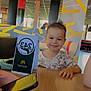 child, toddler, smile, dinosaur_shirt, table, smartphone, hand, restaurant, fast_food, interior, colorful, wall_art, chair, table_number, person, happy, indoor, casual, young, seated
