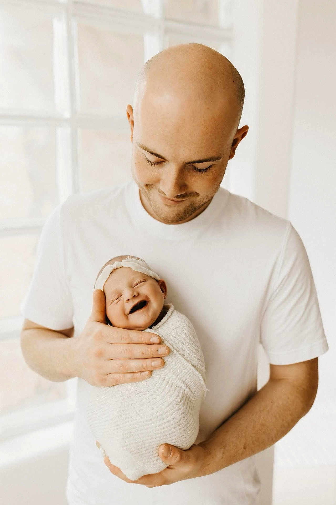 Jacklyn joined the competition — help win amazing prizes! baby, newborn, smile, man, adult, bald, headband, swaddle, white_clothing, window, natural_light, indoor, portrait, father, parent, holding, happy, tenderness, love, family