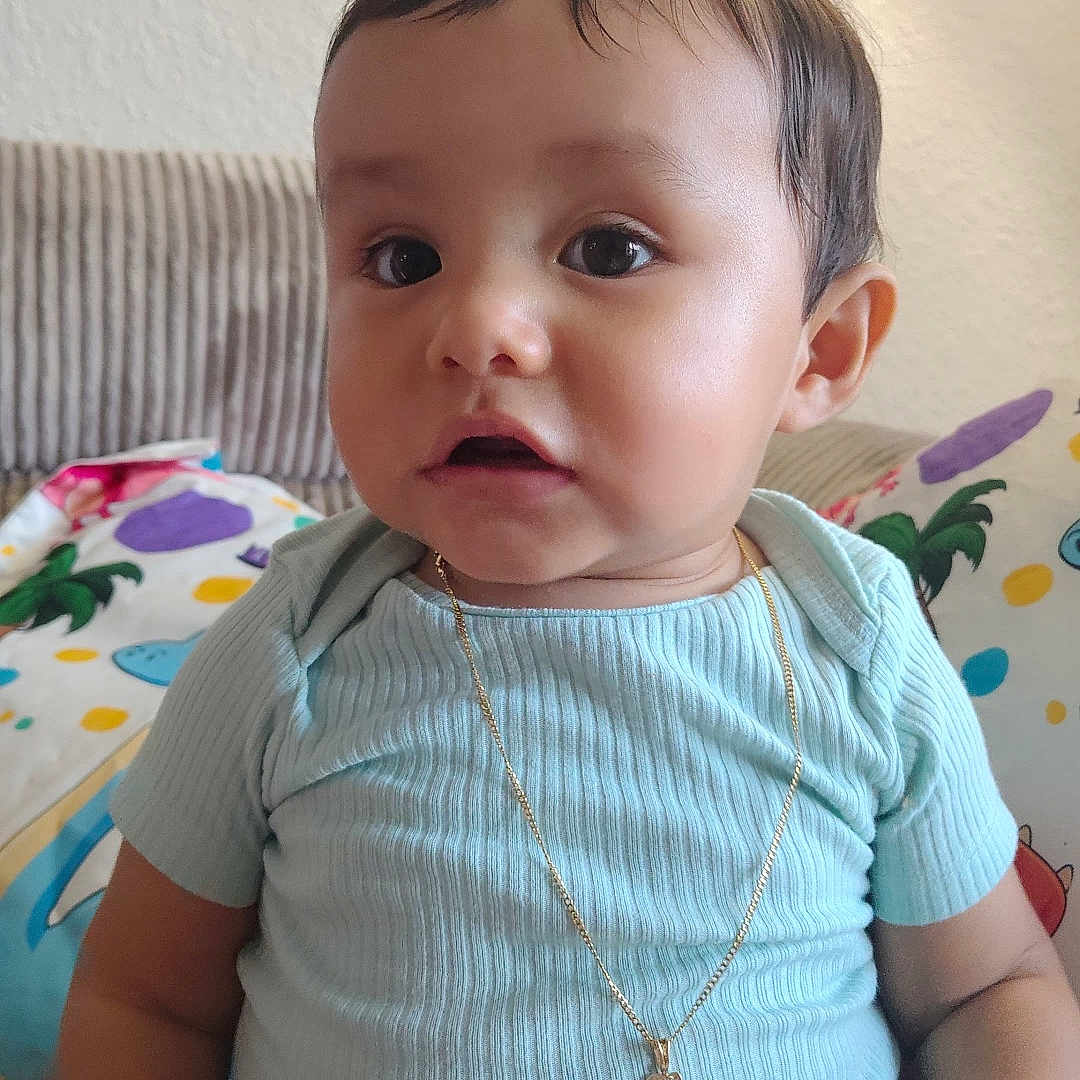 Dylan Frometa joined the competition — help win amazing prizes! baby, child, infant, blue_clothing, necklace, sitting, indoor, couch, blanket, patterned_blanket, curly_hair, face, person, cute, young_child, portrait, hand, home, comfort, expression