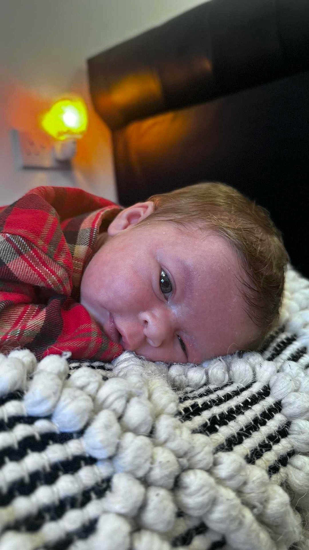 Kylo is registered to the contest to win money with this photo: baby, infant, plaid_shirt, blanket, texture, face, head, sleepy, lying_down, indoor, soft_light, night_light, warm_light, close_up, person, newborn, cozy, resting, bedroom, cute