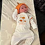 baby, infant, giraffe_pattern, cushion, pumpkin_hat, onesie, cute, child, lying_down, indoors, soft_lighting, white_clothing, orange_hat, patterned_fabric, portrait, small_child, looking_up, baby_clothes, comfortable, resting