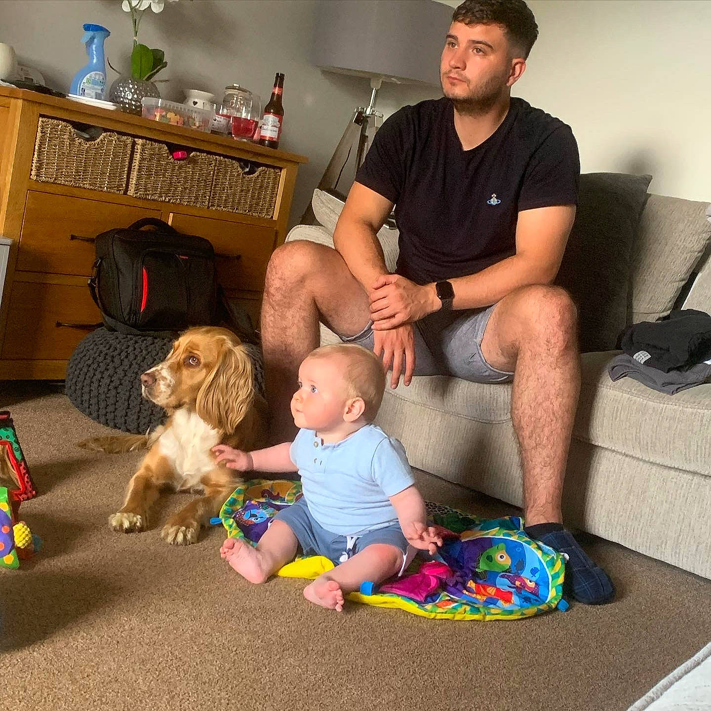 Dougie is registered to the contest to win money with this photo: bookcase, comfort, companion_dog, couch, event, flooring, fun, knee, lamp, lap, leisure, living_room, room, shelf, shorts, sitting, t_shirt, thigh, toddler, toy