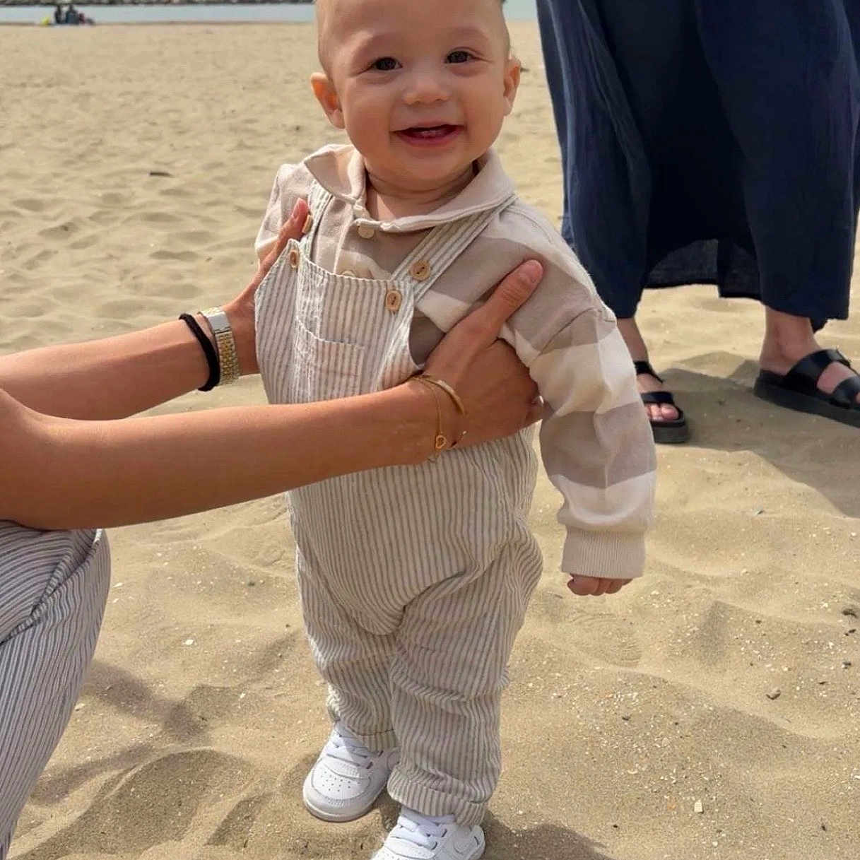 Emilio participe au concours pour gagner de l'argent avec cette photo : child, toddler, beach, sand, smiling, happy, overalls, shoes, adult_hands, person, footwear, dress, sandals, outdoor, sunny, sky, hair, support, casual_clothing, portrait