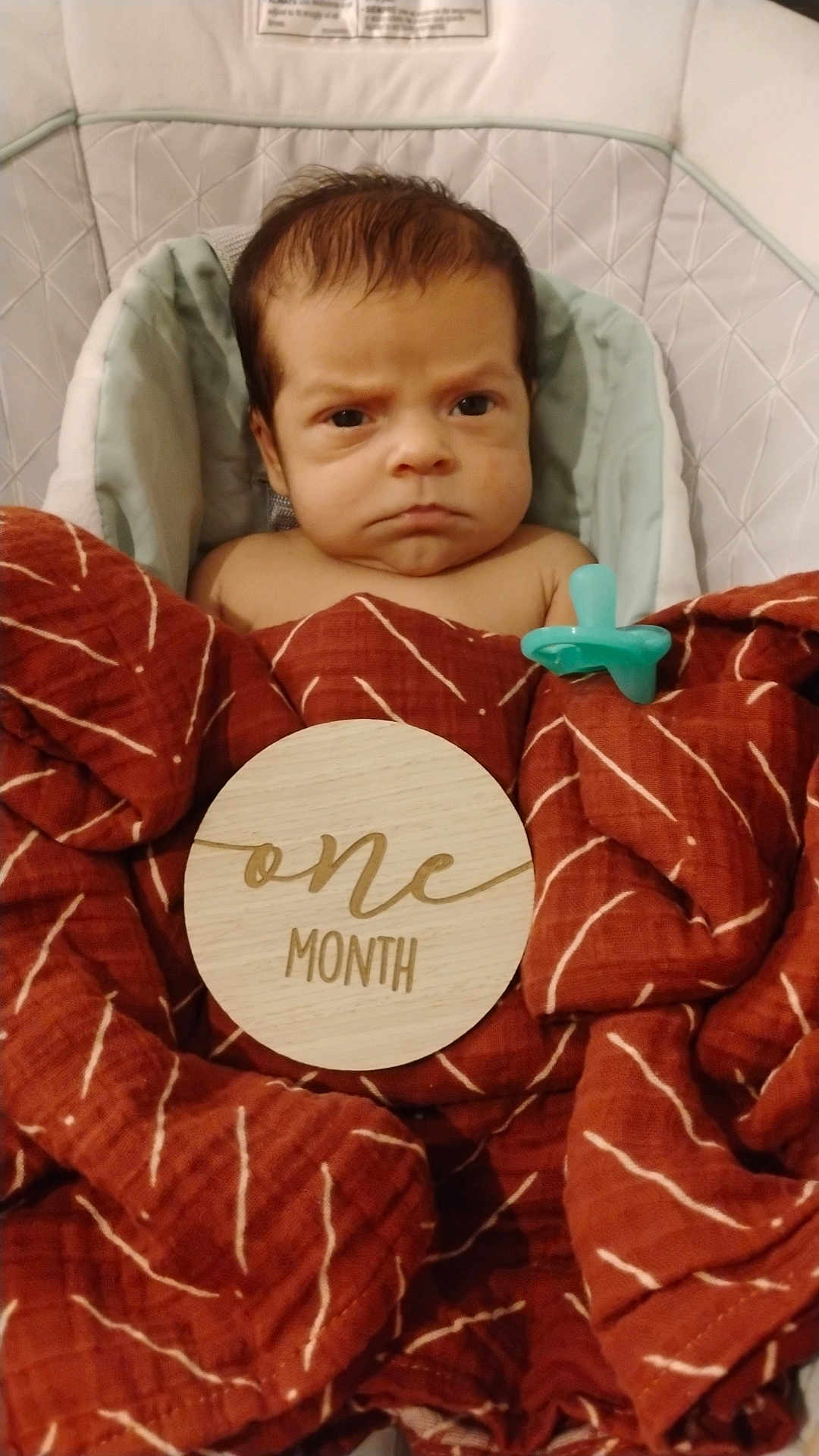 Cooper is registered to the contest to win money with this photo: baby, infant, one_month, blanket, pacifier, baby_seat, face, expression, newborn, wrapped, wooden_sign, text, cushion, clothing, indoor, child, portrait, young, resting, serious