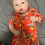 baby, infant, child, orange_clothing, floral_pattern, bracelet, necklace, lying_down, gray_bedspread, blue_eyes, curious_expression, short_hair, cute, indoors, portrait, closeup, soft_lighting, casual_clothing, skin, person