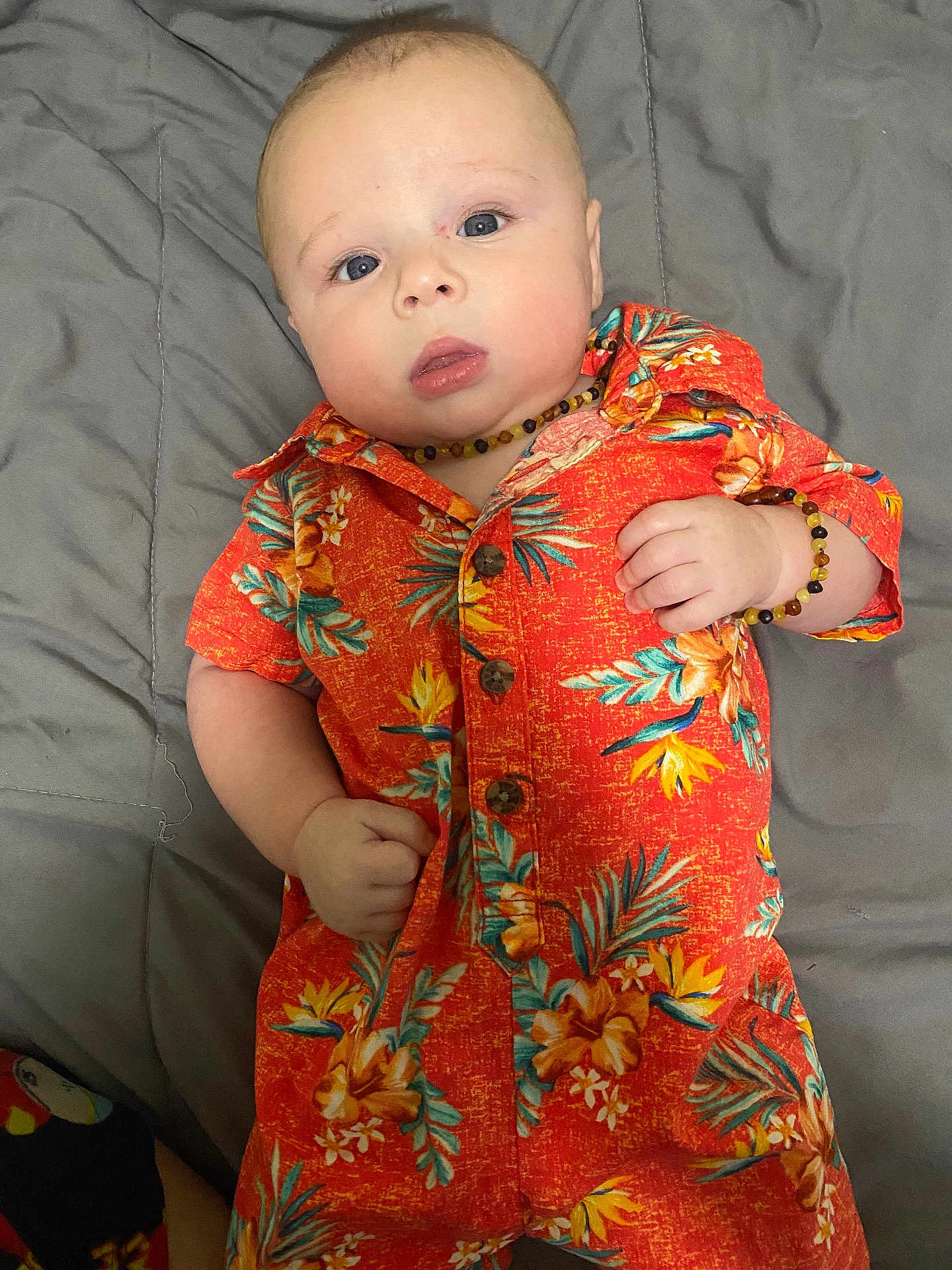 Oliver joined the competition — help win amazing prizes! baby, infant, child, orange_clothing, floral_pattern, bracelet, necklace, lying_down, gray_bedspread, blue_eyes, curious_expression, short_hair, cute, indoors, portrait, closeup, soft_lighting, casual_clothing, skin, person