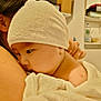 baby, towel, hat, child, person, indoor, home, shelf, wooden_ceiling, skin, face, shoulder, arm, holding, closeup, soft_texture, newborn, resting, warm_lighting, comfort